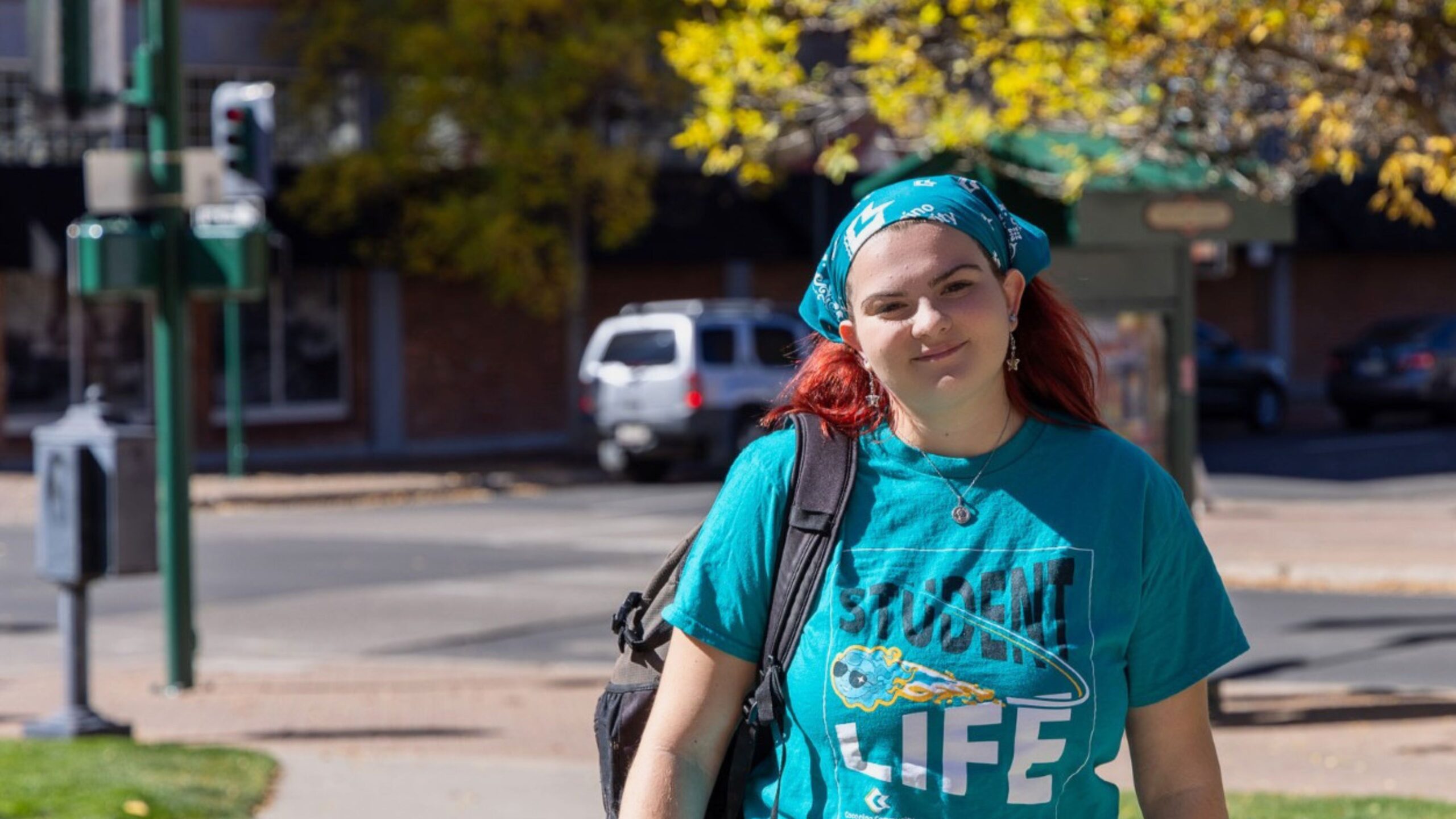 A CCC student wearing a teal Comet Life t-shirt and backpack walks along a campus sidewalk on a sunny fall day, smiling at the camera.